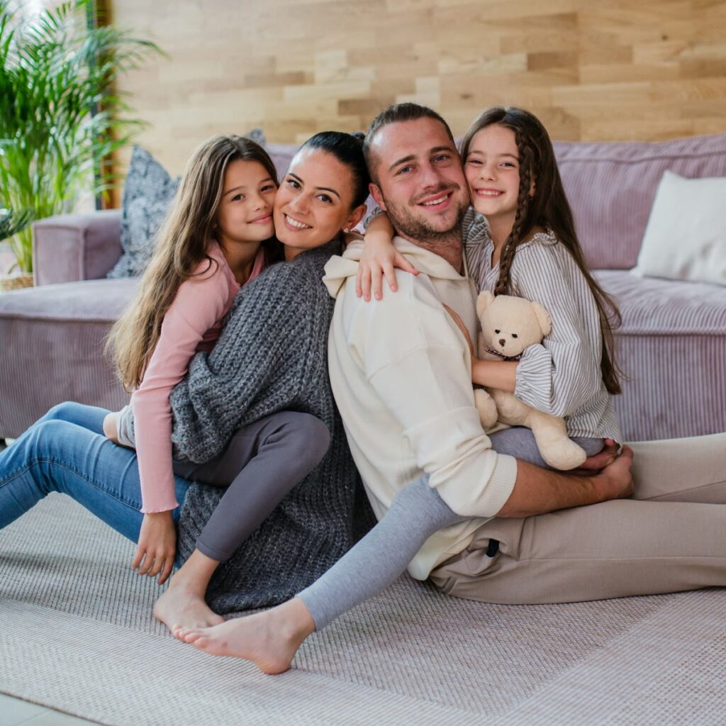 A family of four smiling together on the living-room floor
