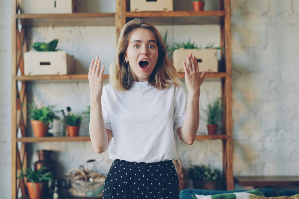 A delighted young woman with her hands raised in a bright living room with plants on wooden shelves