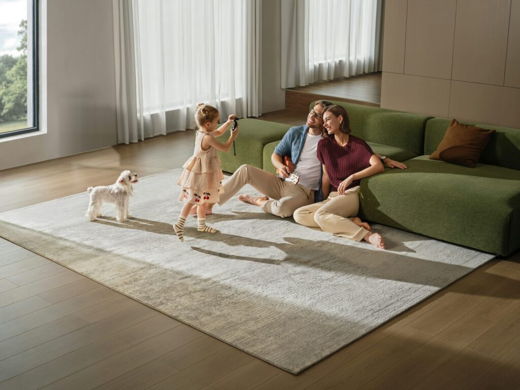 A family laughing together on a sunlit living-room rug while their little girl takes their photo, their dog looking on