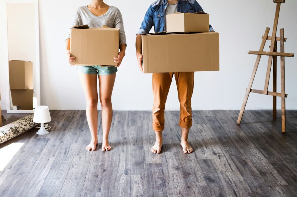 A couple carrying moving boxes into a bright, empty room